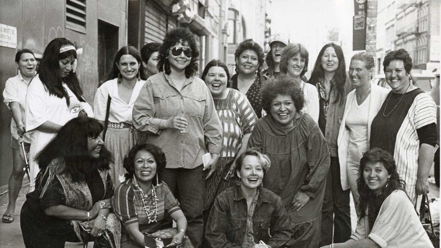 a group of diverse women posing for a photo on sidewalk with buildings in background