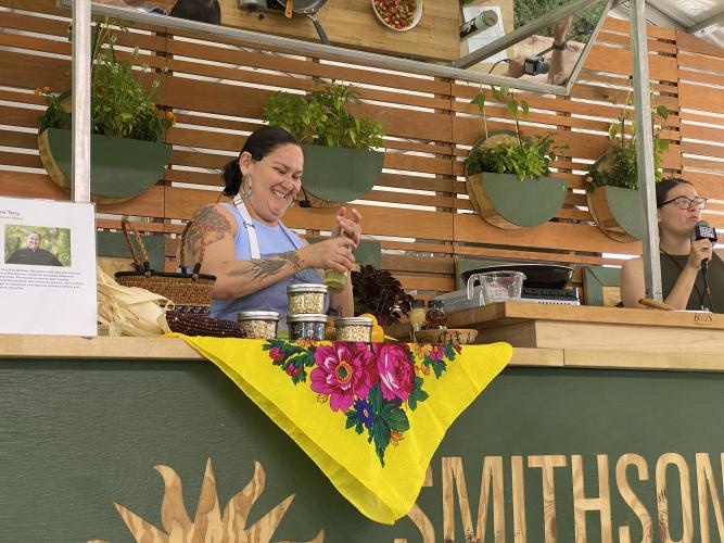 Candid photo of Chef Elena Terry grinding pepper over a dish on the table in front of her. There are four small jars of seeds sitting in front of her. Her daughter Zoe Lea is in the right of the frame holding a microphone.