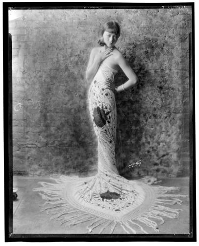 Anna May Wong poses in a full-length, flowered dress that spreads out like a rug at her feet.