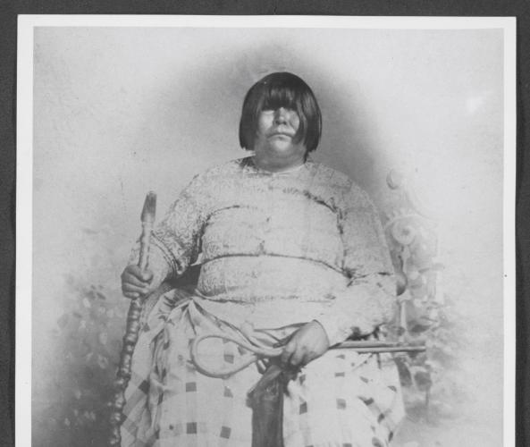 Black and white portrait of Dat So La Lee seated in a chair and surrounded by six decorative baskets on the floor by her feet.