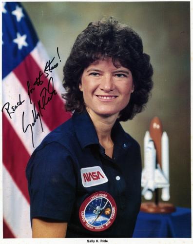 Sally Ride headshot with American flag and a model of a space ship behind her. The photo is signed: Reach for the stars! Sally Ride
