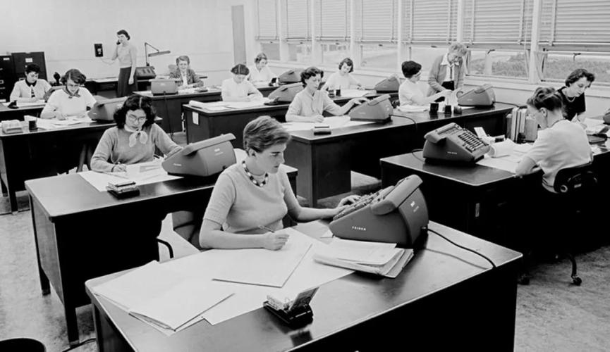 Twelve women sit at two rows of desks with calculators and stacks of papers in front of them. 