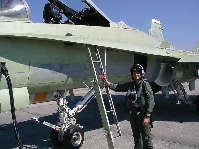 Kim Dyson standing beside a fighter plane preparing to climb up the ladder and get in the cockpit. Her right hand is resting on the ladder, and she is turned looking at the camera.