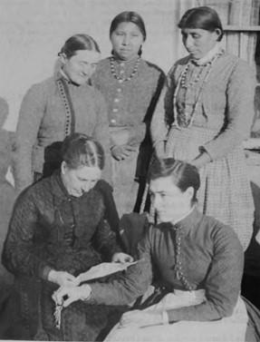 Black and white photo of five women. Three are standing behind two seated in the front. Alice fletcher seated at the bottom left is looking down at a paper in her hands. 
