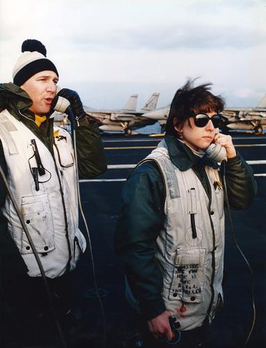 Sharon Deegan standing on the flight deck of the USS Eisenhower holding a phone to her ear. A man slightly behind her is speaking into the phone he is holding. Planes are visible on the deck behind them.