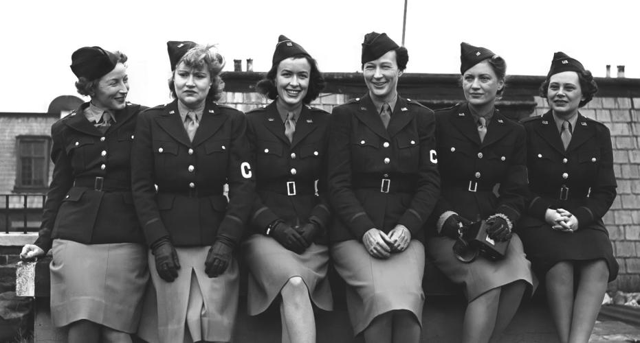 Six women sitting closely together posing for the camera. They are wearing uniforms with light skirts and dark jackets and an armband with the letter C on it.  
