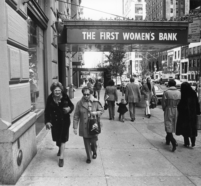 An image of people walking down a street with a sign in the background that reads "THE FIRST WOMAN'S BANK".